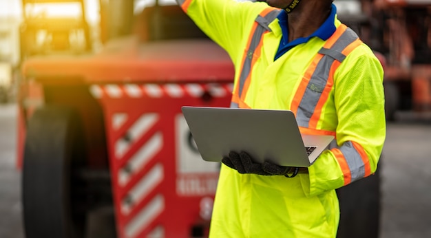 Close up technician dock worker in protective safety jumpsuit uniform and with hardhat and use laptop computer at cargo container shipping warehouse. transportation import,export logistic industrial