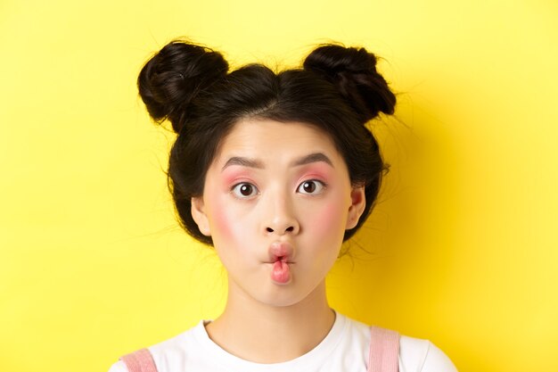 Close-up of teen korean girl pucker lips and looking funny at camera, standing with glamour makeup and stylish hairstyle, yellow.