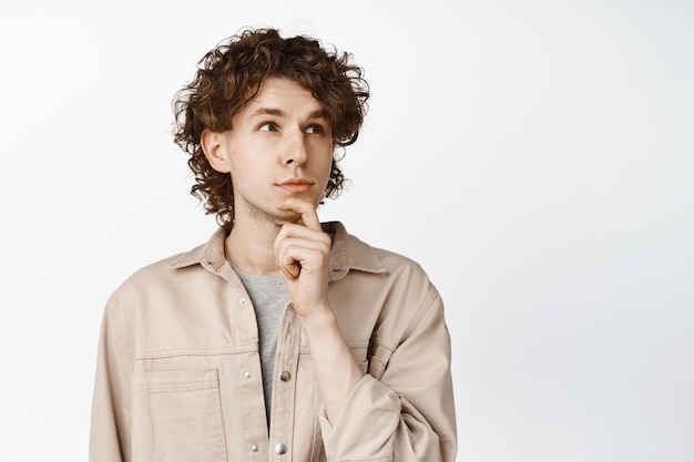 Close up of thoughtful male student touch his chin and ponder looking up thinking while standing over white background