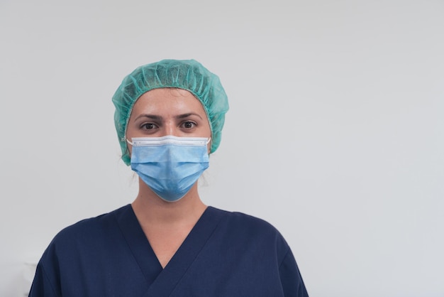 Close up of tired female doctor or scientist with a medical mask and surgical cap over grey background. Authentic real medical people hero in white in coronavirus pandemic is adjusting mask.