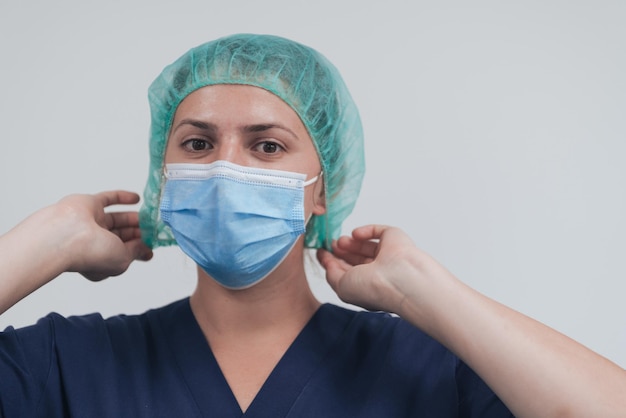 Close up of tired female doctor or scientist with a medical mask and surgical cap over grey background. Authentic real medical people hero in white in coronavirus pandemic is adjusting mask.