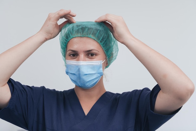 Close up of tired female doctor or scientist with a medical mask and surgical cap over grey background. Authentic real medical people hero in white in coronavirus pandemic is adjusting mask.