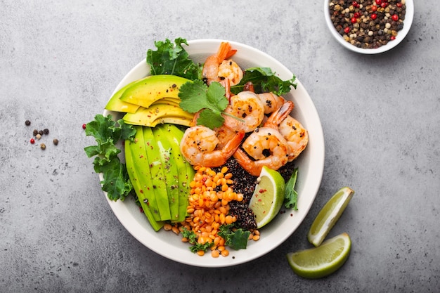 Close-up, top view of salad bowl with shrimps, avocado, fresh kale, quinoa, red lentils, lime and olive oil on gray stone background. Lunch bowl, healthy clean eating, dieting or nutrition concept