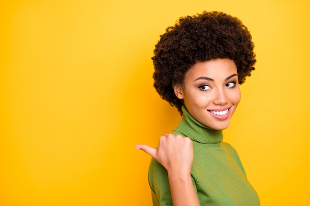 Close up turned portrait of cheerful positive nice trendy woman pointing into empty space with her thumb smiling toothily .