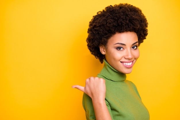 Close up turned portrait of curly wavy charming woman pointing into empty space behind her back with thumb .