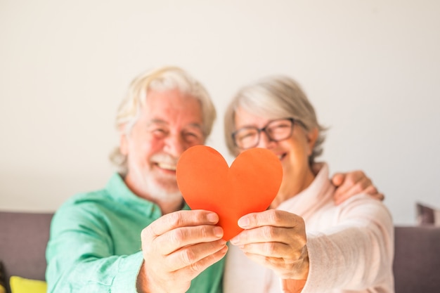 Close up of two happy and in love seniors smiling and looking at the camera holding a red heart together - mature people in love