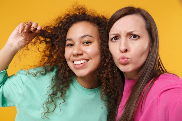 Close up of two women friend european african american girl in pink green clothes posing isolated on yellow background. People lifestyle concept. Mock up copy space. Doing selfie shot on mobile phone.