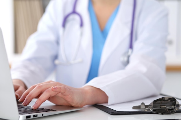 Close up of unknown female doctor typing on laptop computer while sitting  at the table. Medical staff, compulsory work concept