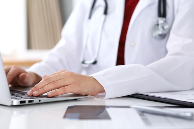 Close up of unknown female doctor typing on laptop computer while sitting at the table