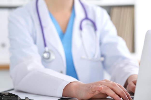 Close up of unknown female doctor typing on laptop computer while sitting at the table