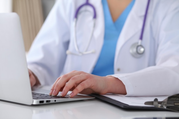 Close up of unknown female doctor typing on laptop computer while sitting at the table