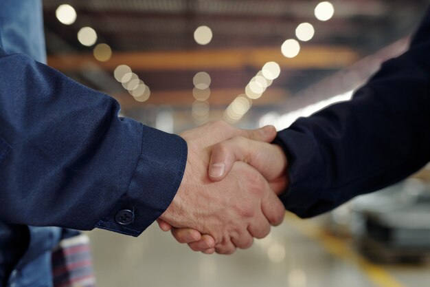 Close-up of unrecognizable factory executives making handshake in industrial shop with lights