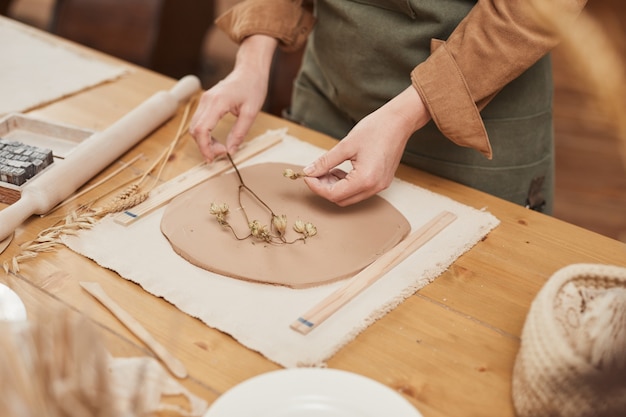 Close up of unrecognizable female artisan making plant imprint ceramics on wooden table in pottery workshop, copy space