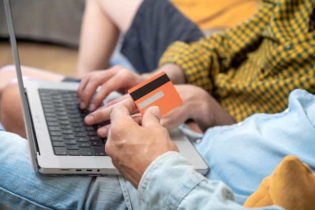 Close up of an unrecognizable gay couple using laptop computer while sitting on a couch at home shopping online with a credit card