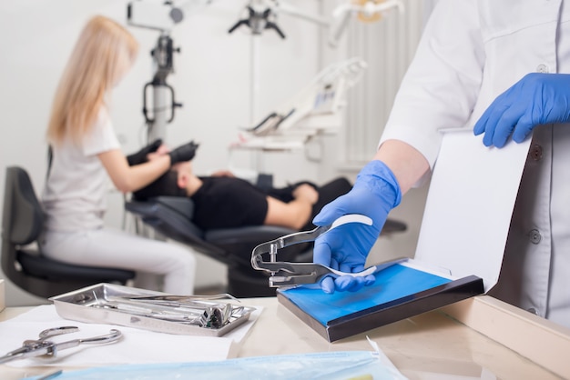 Photo close-up view of assistant's hands with blue gloves working with dental tools, on the blurred background dentist is treating patient in dental clinic. dentistry