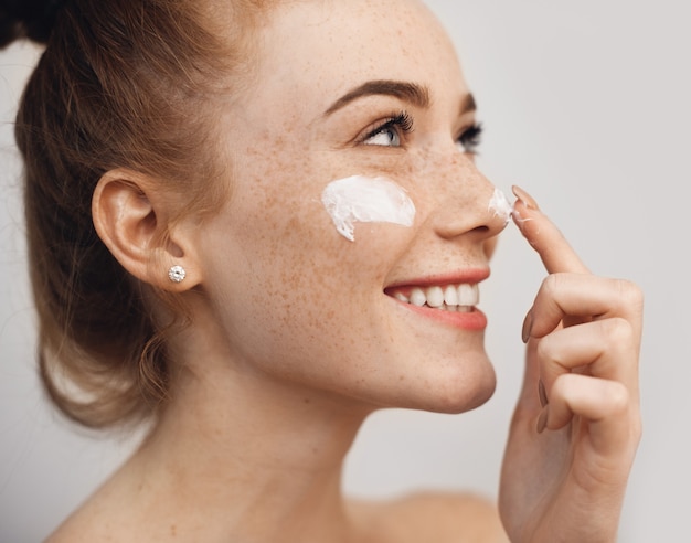 Close up view of caucasian red haired woman with freckles applying a cream on her face while posing aside on a whole wall with freespace