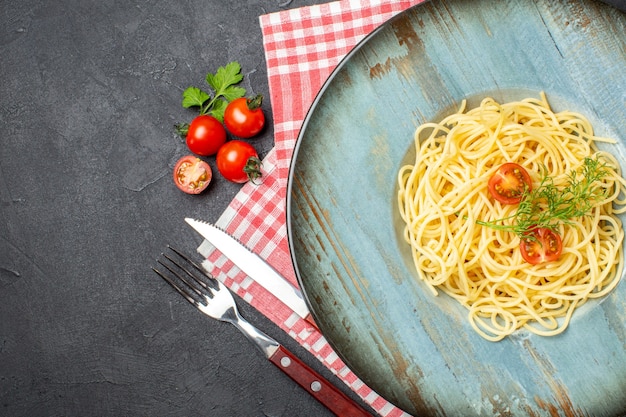 Close up view of delicious spagetti served with tomatoes greens and cutlery set on red stripped towel on the left side on black background