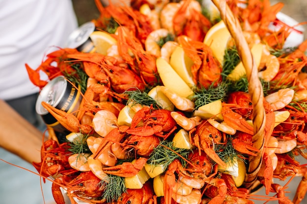 Close-up view over a festive basket full of fresh and tasty crayfish, shrimps and beer cans decorated with sliced lemon and bunches of dill.