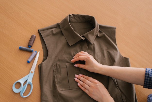 Close up view from above on the hands of a dressmaker a young woman is manually working on the design of a new collection of khaki shirts There are threads and scissors on the table nearby