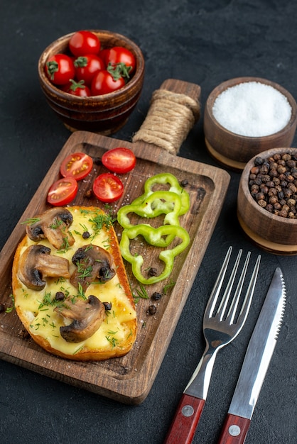 Close up view of homemade tasty snack with mushrooms and chopped vegetables on wooden board cutlery set spices on black background