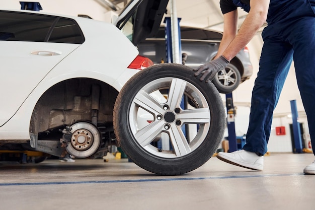 Close up view of man in work uniform with car wheel indoors Conception of automobile service