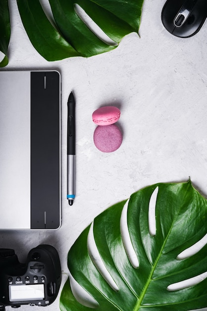 Close-up view of photographer's of graphic designer's workplace. Tablet, stylus, camera and zoom lens, optical wireless mouse, macarons, monstera green leaf.