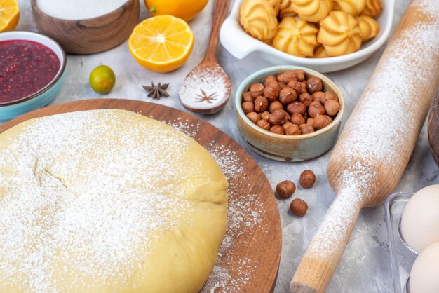 Close up view of raw pastry flour on round board grater fresh fruits jam cookies hazelnuts on stained white background