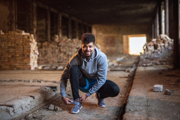 Close up view of sporty active afro american runner man crouching and tying velcro sneakers in the abandon place.