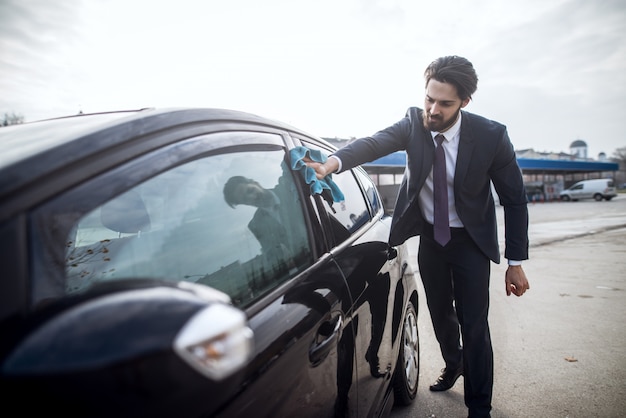 Close up view of stylish bearded handsome young hardworking man in suit cleaning his black car with a blue microfiber cloth on the manual self-service car washing station.