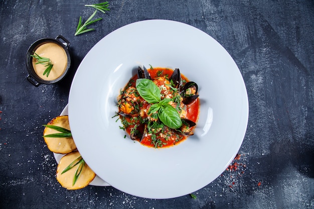 Close up view on tasty seafood soup in white restaurant bowl. bouillabaisse with mussels, salmon and basil with a snack of potatoes with cheese sauce. Dark background. Food photo for menu or recipe