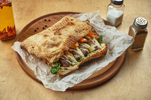 Close up view on tasty traditional italian sandwich panuozzo - oven-baked tortilla with tomatoes, melted mozzarella, minced beef and arugula on a wooden board. Street food