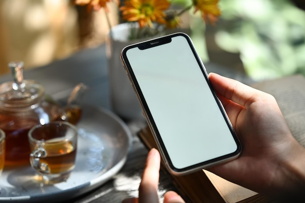 Photo close up view of woman hand holding smart phone with traditional tea set and flower pot on background