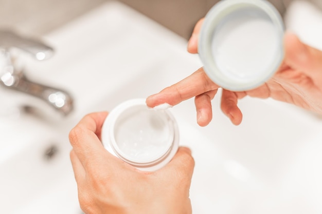 Close-up view of a woman's hands holding a pot of moisturizing cream and taking a fingerful of cream for application. Concept of skin care.