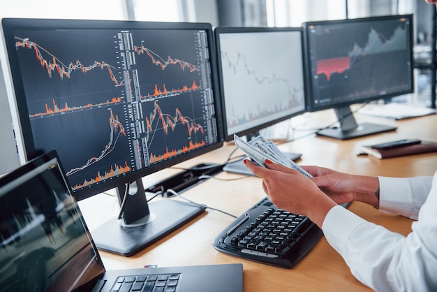 Close up view of woman's hands that holds money near the monitors with graphs.