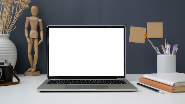 Close up view of workspace with open blank screen laptop, painting tools and office supplies on white table with grey wall