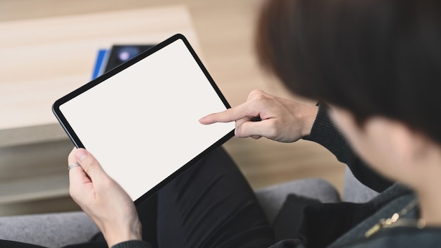 Close up view of young man holding horizontal digital tablet with blank screen while sitting on couch.