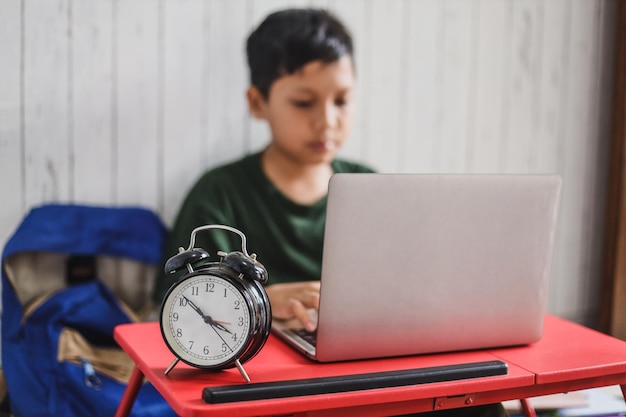 Close up vintage an alarm clock stands on the school desk with blurred asian boy in online learning