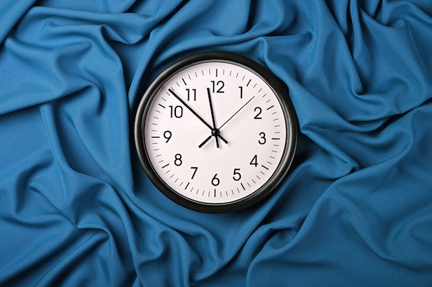 Close up white wall classic clock over blue textile background with folded pleats of fabric, elevated top view, directly above