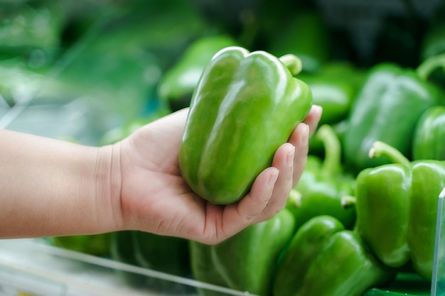 Close up woman hand holding fresh green bell pepper on the shelf at supermarket