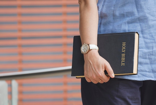 Close up woman hand holding holy bible while standing with blurred background in public area