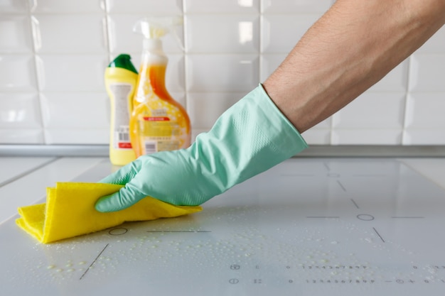 Close up of woman hand in protective rubber gloves washing or cleaning modern white induction hob by a rag in the kitchen, blurred cleaning products