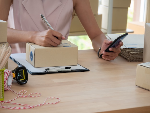 Close up of woman hand seller writing address on parcel box and checking product order on smartphone