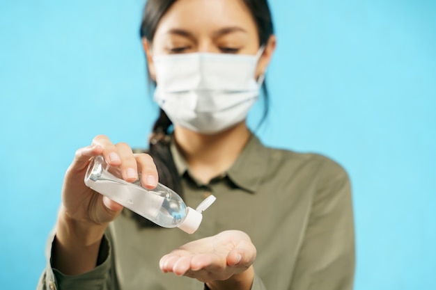 Close up of woman hands in medical protective mask using gel sanitizer on blue background