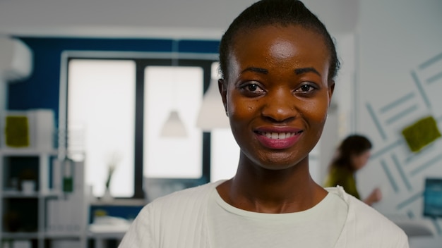 Close up of woman looking at camera smiling standing in start up creative agency office holding laptop, typing on it