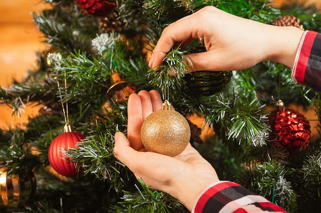 Close-up of a woman in a plaid shirt hangs a beautiful shiny gold ball on a Christmas tree