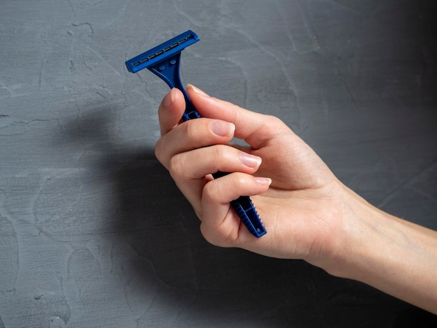 Close up of a woman's hand holding a blue plastic razor on a gray textured background