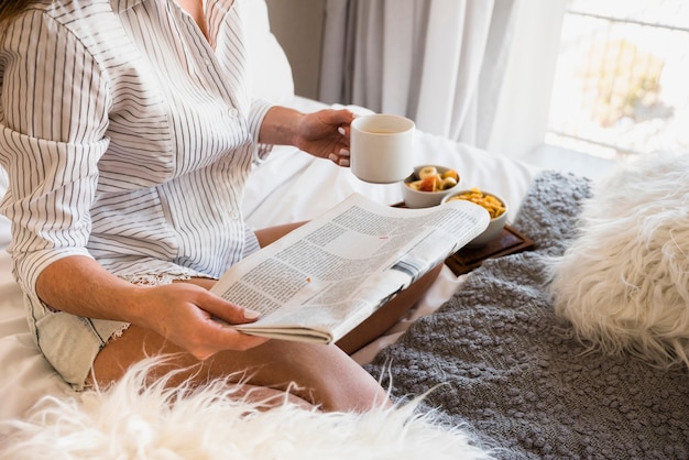 Close-up of a woman sitting on bed with newspaper and coffee cup in hand