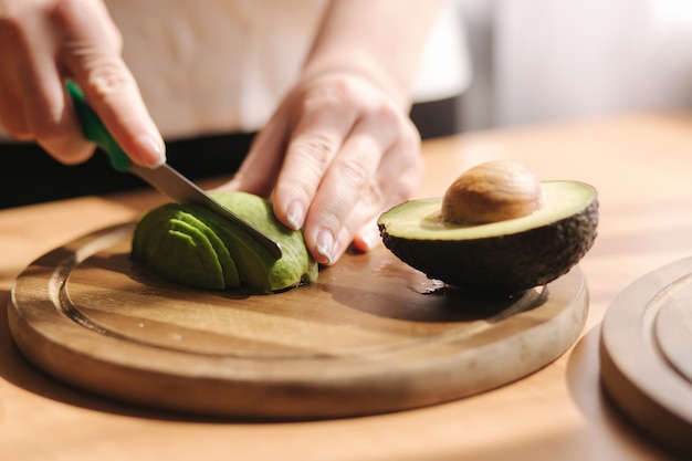 Close up of woman slicing avocado on wooden board at home. Vegetarian food concept