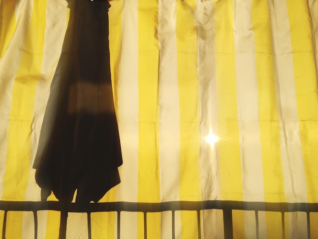 Photo close-up of yellow curtain with sunlight peaking through and the silhouette of a closed umbrella