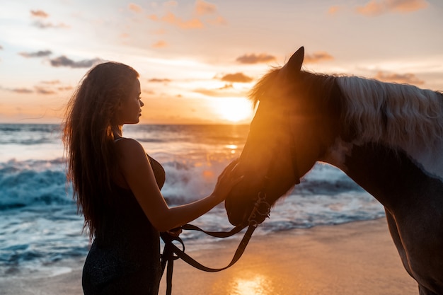 Close up: Young brunette beauty having fun with horse on the beach. Sunset